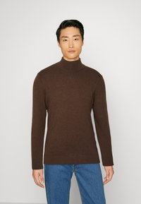 Mørkebrun rullekravegenser laget av et mykt strikkemateriale, med lange ermer og en tettsittende silhuett, kombinert med blå denimjeans.