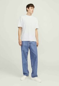 White cotton t-shirt with a crew neck, paired with light blue denim jeans. Model wears white slip-on shoes, standing against a neutral background.