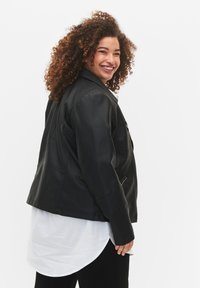 Veste en cuir noir au design structuré, dotée d'un col à revers et de poches zippées, portée avec une longue chemise blanche en dessous.