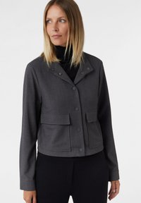 Kobieta ubrana w ciemnoszary, zapinany na guziki płaszcz z klapkowymi kieszeniami na czarnym golfie, spoglądająca w bok na tle gładkiej powierzchni.