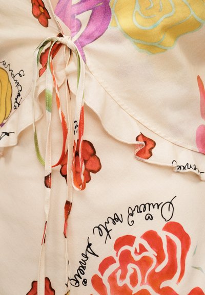 Κοντινό πλάνο μπεζ υφάσματος με πολύχρωμα λουλουδένια σχέδια, φραμπαλά στην άκρη και λεπτές δεμένες τιράντες σε κόκκινους, πράσινους και μπεζ τόνους.