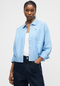 Γυναίκα με πλεξούδα φορά μπλε ριγέ πουκάμισο με κουμπιά πάνω από λευκό τοπ, μαύρο παντελόνι, με το ένα χέρι στην τσέπη, απέναντι σε απλό φόντο.