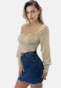 Beiges Langarmshirt mit gerafften Details an der Brust, kombiniert mit einem hoch taillierten dunklen Jeansrock mit ausgefranstem Saum und Taschen.