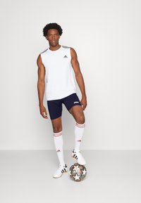 Maglietta atletica bianca senza maniche, pantaloni corti blu scuro con logo, calze a righe rosse e bianche, scarpe da ginnastica bianche e un pallone da calcio con accenti neri e arancioni.