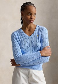 Žena s pletenými vlasmi, oblečená v svetlomodrom svetríku s vrkočovým vzorom a výstrihom do V a bielych nohaviciach, so založenými rukami, pozerá sa do strany.