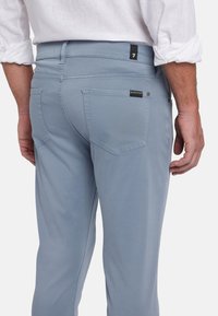 Lichtblauwe broek van zacht materiaal, met een achterzak met een accentlabel en gestikte details, zijkant weergegeven.