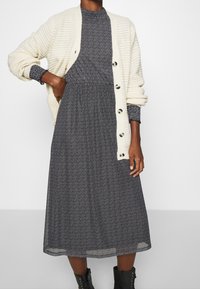 Kremowy kardigan ze żebrowanym wzorem, zapinany na duże guziki, noszony na czarnej sukience w białe kropki, z rozkloszowaną spódnicą do kostek.