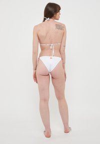 Set bikini bianco con collo a halter e chiusura con lacci. Presenta un design a copertura minima con slip decorati da dettagli rosa e un'etichetta con logo sul retro.