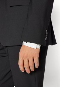 Schwarze Anzugjacke aus strukturiertem Stoff, mit sichtbarem weißen Hemdmanschettenmuster in Schwarz. Hand liegt an der Seite.