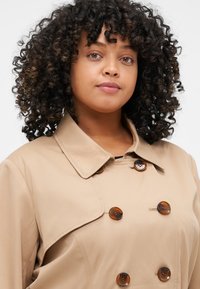 Beige trenchcoat med dobbeltradet design, store brune knapper og klassisk krage. Glatt tekstur og skreddersydd passform.