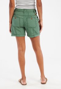 Groene katoenen shorts met opgerolde zoom, elastische tailleband en twee achterzakken met bruine knopen. Gestreept shirt is gedeeltelijk zichtbaar.