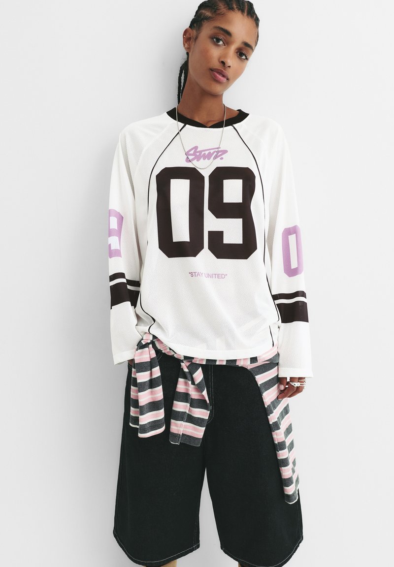 Weißes Mesh-Hemd mit langen Ärmeln und schwarzen sowie rosafarbenen Akzenten, mit dem Text "09" und "STAY UNITED". Lockere schwarze Shorts; gestreifte Krawattenbluse.