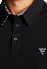 Acercamiento de camiseta polo negra con ribete gris en el cuello, dos botones y logo triangular gris de Guess en el pecho.