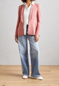 Růžový blazer s klopami, zkombinovaný s bílou košilí na knoflíky a světle modrými širokými džíny. Bílí tenisky dotvářejí outfit.