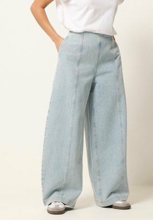 Personne portant un jean large bleu clair avec des coutures verticales, un t-shirt blanc rentré, les mains dans les poches et des baskets blanches.