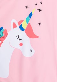 Un tissu rose présente une illustration de licorne blanche avec une corne multicolore, les yeux fermés et une crinière rouge ; des accents volants bleus et verts sur le côté.