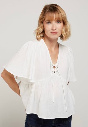 Femme aux cheveux blonds courts portant un chemisier blanc ample texturé avec de larges manches volantes et un col en V à lacets, debout devant un fond uni.