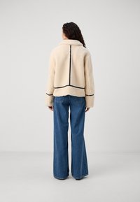 Cream-colored fleece jacket with a black trim, featuring a wide collar and back seam detailing. Paired with blue wide-leg jeans.