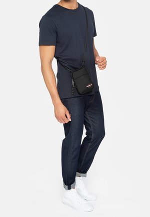 Man draagt een navyblauw T-shirt en donkere jeans met opgerolde pijpen, witte sneakers en een zwarte crossbodytas met logo aan de voorkant.