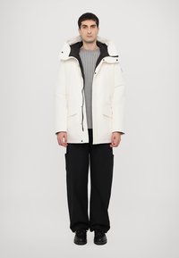 Parka blanche et isolée avec un col haut, fermeture éclair à l'avant et poches latérales doubles. Portée par-dessus un pull en laine gris côtelé et un pantalon cargo noir.