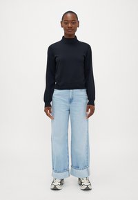 Jūras zilas turtleneck džemperis ar garām piedurknēm, apvienots ar gaiši ziliem platām kājām džinsiem, salocītām piedurknēm un rakstainām kedām. Vienkāršs, mūsdienīgs dizains.