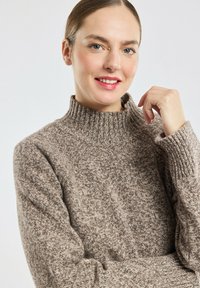 Jeune femme à la peau claire et aux yeux bleus, portant un pull en tricot marron texturé avec un col montant à côtes, souriant doucement, main près de son visage.