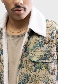 Giacca a fantasia floreale realizzata in tessuto testurizzato con colletto in pile bianco. Presenta una tasca sul petto e una camicia beige a strati sotto.