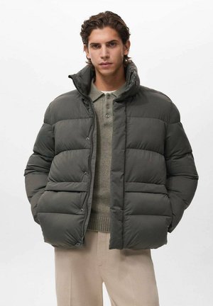 Chaqueta acolchada gris con cuello alto, cierre de botones y diseño acolchado horizontal. Usada sobre una camisa de polo gris claro.