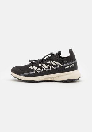 Zapatilla de trail running Adidas Terrex en blanco y negro con talón acolchado, puntera reforzada y sistema de cordones rápidos sobre fondo blanco.