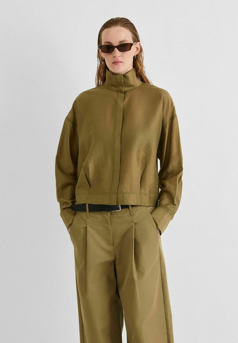 Personne portant une veste col montant vert olive, un pantalon large assorti, une ceinture noire et des lunettes de soleil sombres, debout les mains dans les poches.