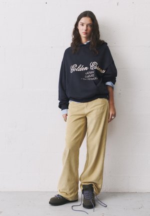 Tengerészgyümölcs színű kapucnis pulóver "Arany Esszencia" logóval, széles, világossárga kordbársony nadrággal párosítva. Sötét fűzős csizma lila cipőfűzőkkel.