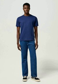 Σκούρο μπλε κοντομάνικο μπλουζάκι συνδυασμένο με ίσια μπλε τζιν παντελόνια και μαύρα αθλητικά παπούτσια με λευκή σόλα, σε φωτεινό φόντο.