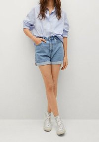 Mujer vestida con una camisa de botones a rayas azul claro, pantalones cortos de mezclilla enrollados y zapatillas altas blancas, de pie con una mano en el bolsillo.