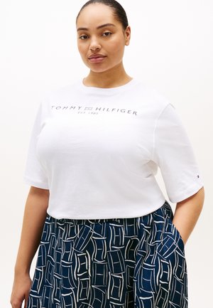 Ženska nosi belo majico Tommy Hilfiger z logotipom, kombinirano s temno modro krilo z belim abstraktnim geometrijskim vzorcem.