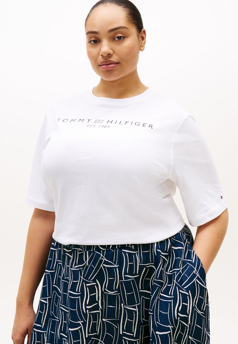 Γυναίκα φοράει λευκό T-shirt Tommy Hilfiger με κείμενο λογότυπου, συνδυασμένο με μια σκούρα μπλε φούστα με άσπρο αφηρημένο γεωμετρικό σχέδιο.