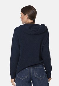 Felpa blu navy realizzata in tessuto morbido e testurizzato, caratterizzata da una vestibilità rilassata, cappuccio profondo e polsini a coste, abbinata a jeans in denim strappati.