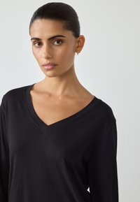 Czarna bluzka z długim rękawem i dekoltem w kształcie litery V, wykonana z miękkiego materiału. Modelka ma gładkie włosy i nosi małe kolczyki koła.