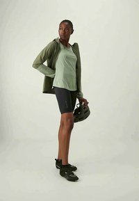 Αθλητικό άτομο με πράσινο μπουφάν, πράσινη μπλούζα, μαύρες ποδηλατικές βερμούδες, κρατώντας ένα μαύρο κράνος ποδηλάτου, όρθιο σε άσπρο φόντο.