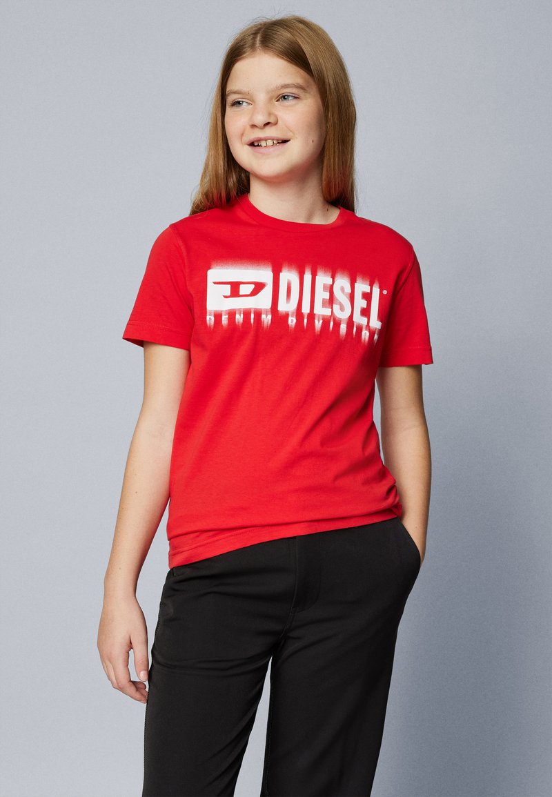 Rød bomulls t-skjorte med en hvit "DIESEL" logo med gradienteffekt. Rund hals og korte ermer. Kombinert med svarte bukser.