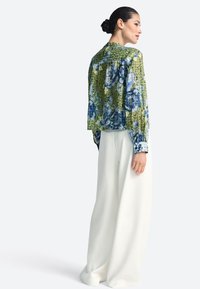 Bluză cu model floral verde și albastru, cu o siluetă lejeră, purtată peste pantaloni albi cu picioare largi, având mâneci cu manșete și un design fronsat în partea din spate.