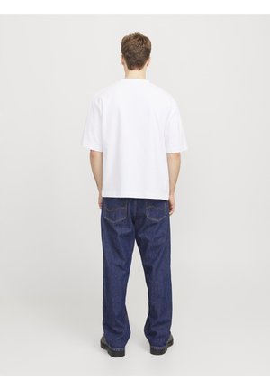 Mann steht mit dem Rücken zur Kamera, trägt ein weißes kurzärmeliges Hemd, blaue Jeans und schwarze Schuhe vor einem schlichten hellen Hintergrund.