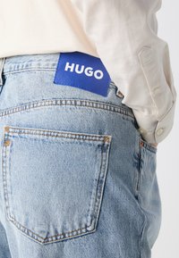 lichtblauwe denim jeans met een opvallende blauwe leren patch waarop "HUGO" in het wit is gedrukt. Bevat een achterzak met oranje stiksel.