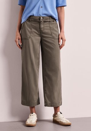 Person iført olivengrønne, brede, korte bukser, lyseblå kortærmet skjorte og hvide sneakers, stående mod en lys væg.