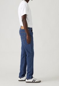 Homme debout en jean bleu, t-shirt blanc et baskets blanches avec des rayures noires sur un fond clair uni.
