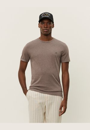 Braunes Kurzarmshirt mit Rundhalsausschnitt und kleinem Logo auf der Brust, kombiniert mit hell gestreiften Hosen und einer schwarzen Kappe.