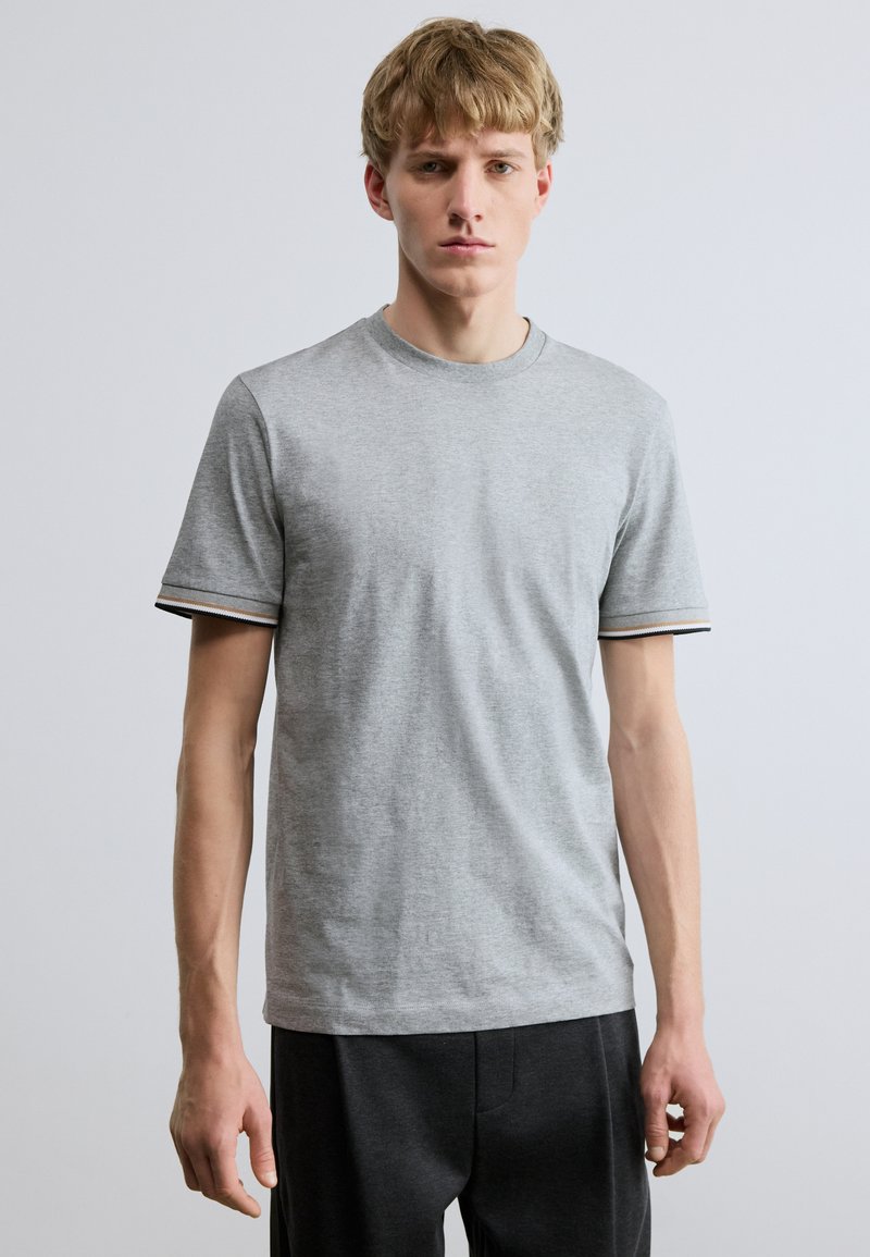 Szürke pamut póló kerek nyakkivágással és rövid ujjakkal. Fekete-fehér csíkos részletek találhatók az ujjvégeken. Egyszerű, testhezálló fazon.