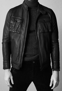 Homme portant une veste en cuir noire sur un pull à col roulé en maille torsadée foncé avec un pantalon noir, debout les mains le long du corps.