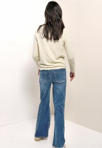 Femme aux longs cheveux bruns, vêtue d'un pull torsadé crème et d'un jean large bleu, debout dos à la caméra devant un mur uni.