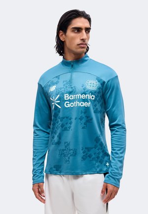 Homme portant un haut de sport bleu à manches longues avec l'emblème "Bayer Leverkusen" et les logos des sponsors "Barmenia Gothaer", assorti à un short blanc.