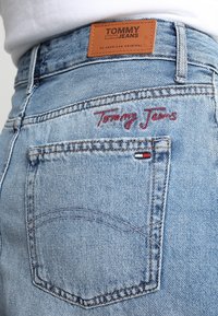 Blå denimjeans med en läderetikett som har "TOMMY JEANS" och röd broderad text. Detaljerad sömnad och en liten logotypdetalj på fickan.